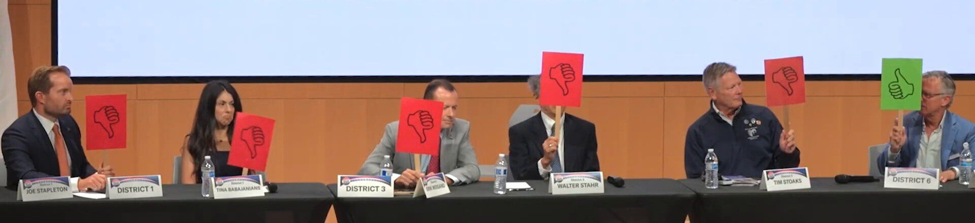 Six Newport Beach council candidates at a table holding up cards. Five cards are red thumbs-down. The candidate at the far right, Andrew Gerken, holds a green thumbs-up.